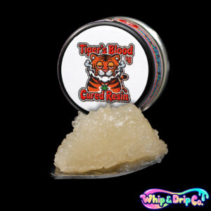 “Whip & Drip Co. Tigers Blood cured resin concentrate with golden extract displayed beside colourful tropical-themed 1 gram product packaging on a black background.”